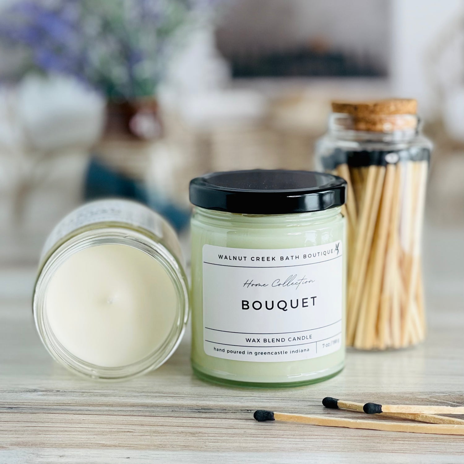 bouquet candle with lid to the right of another candle with the top off to show the creamy wax inside. there are a few matches scattered in front and a pretty glass jar of matches in the background
