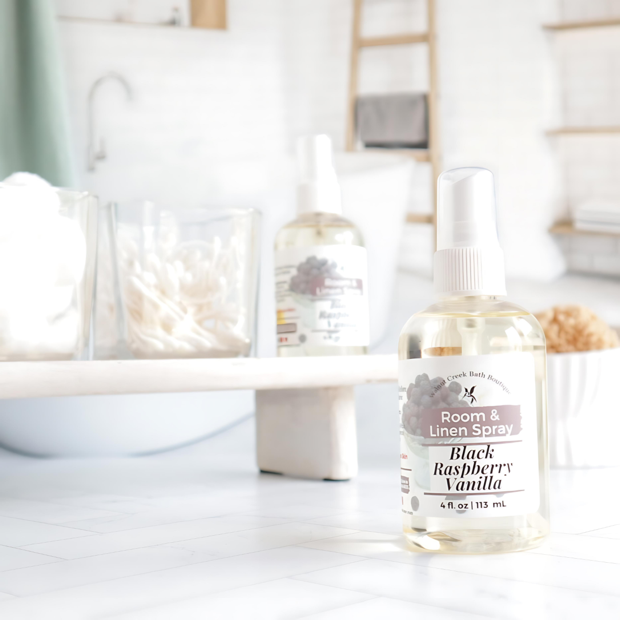 black raspberry vanilla room and linen spray is front right, behind this is a try with cotton balls and qtips in glass jars with another room spray next to them. in the background is an image of a tub and shelf in a spa like bathroom