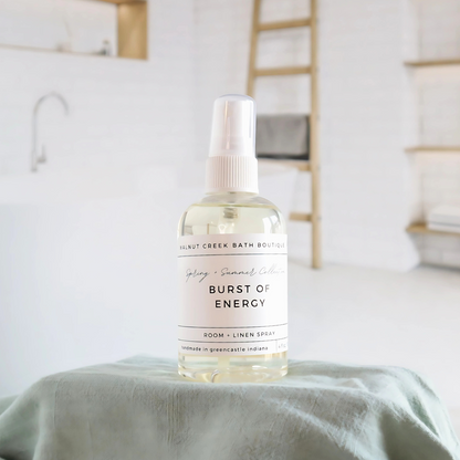 burst of energy room and linen spray is in a clear bottle with a white spray nozzle. it is sitting on a raised platform with a green towel unter it and in the background is a spa like bathtub