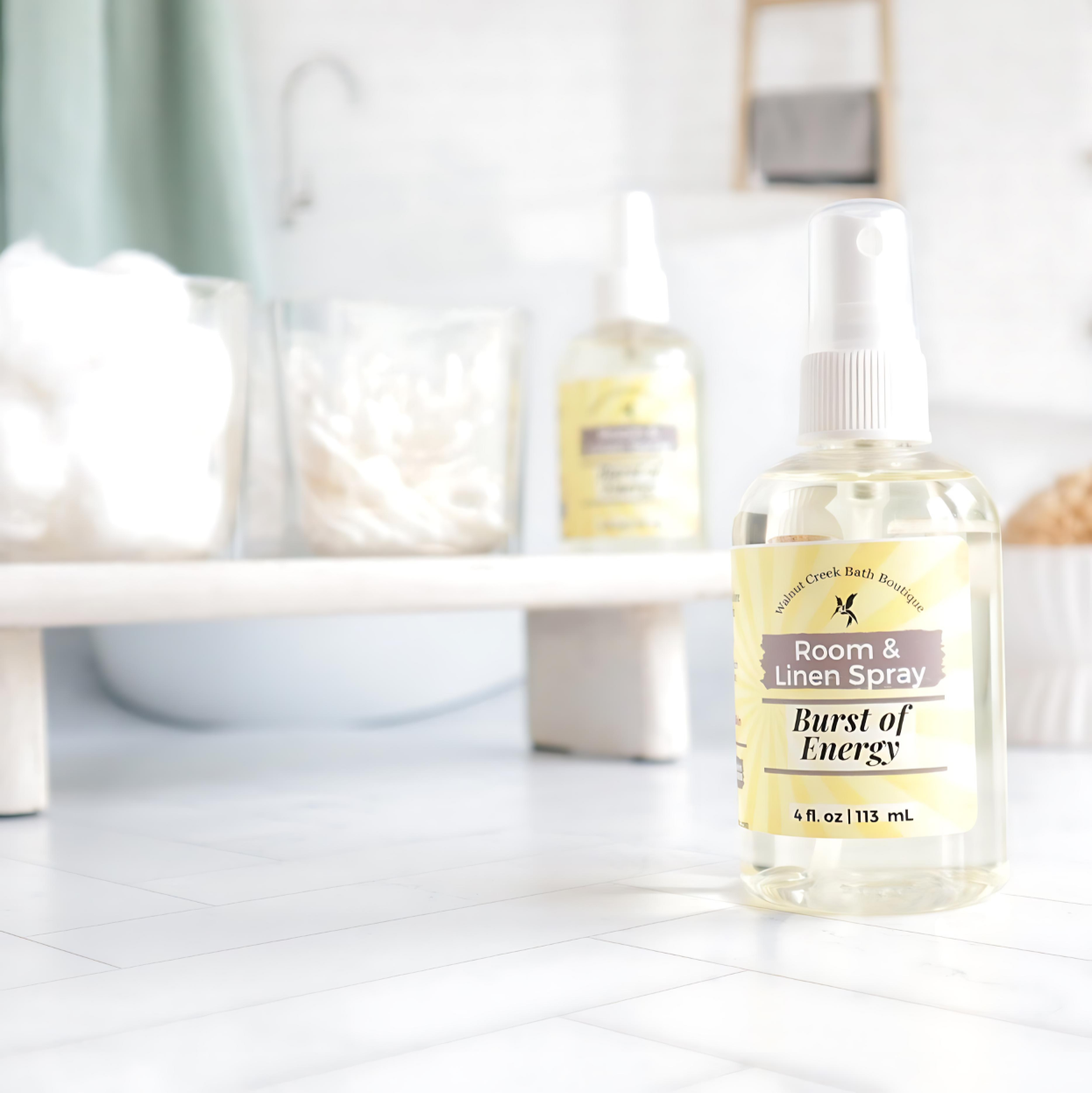 burst of energy room and linen spray is front right, behind this is a try with cotton balls and qtips in glass jars with another room spray next to them. in the background is an image of a tub and shelf in a spa like bathroom