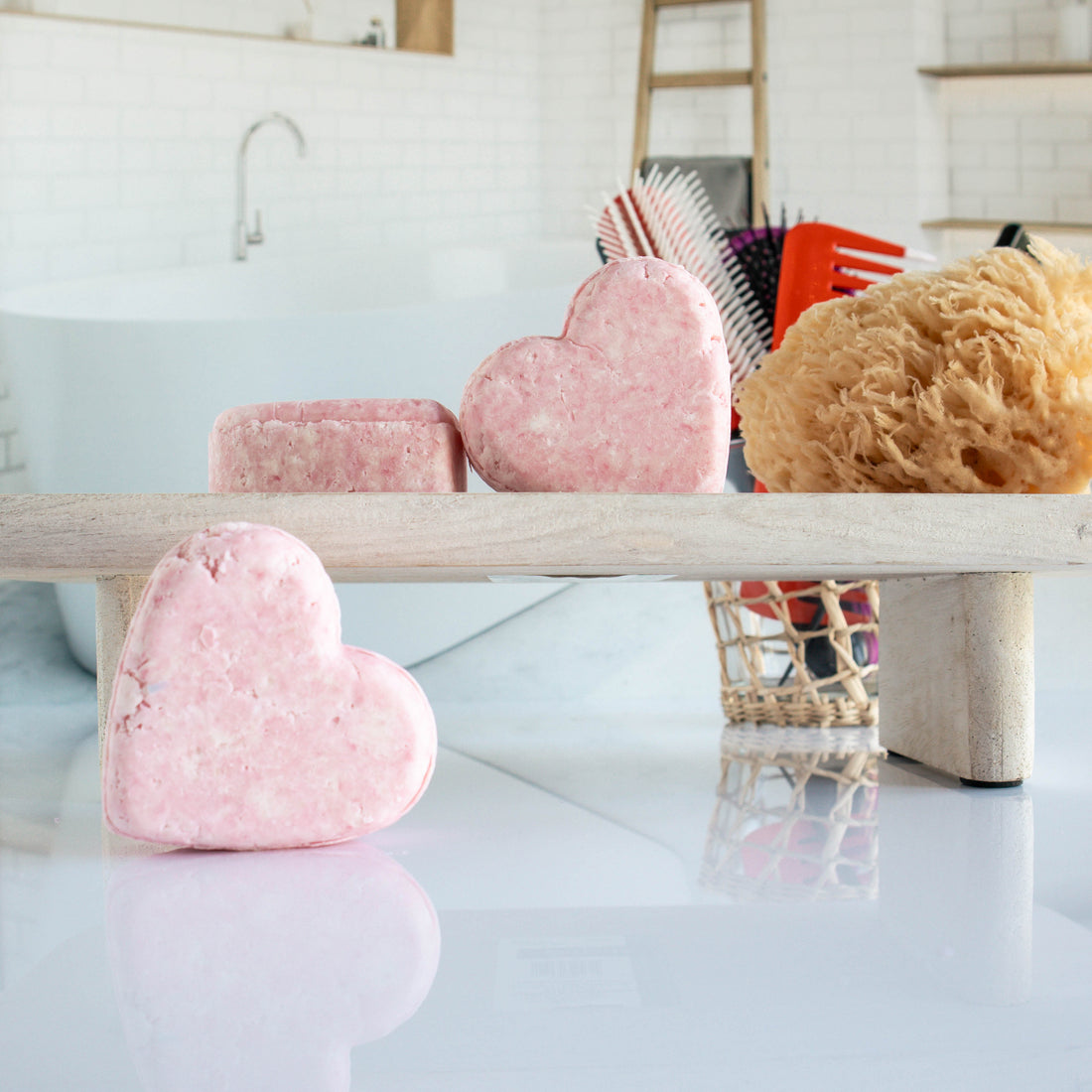 3 pink burst shampoo bars shaped in a heart. One is leaning on its side while the other two are on a trey, one leaning on the other. there is a loofah next to them with a cup of hair tools in the background