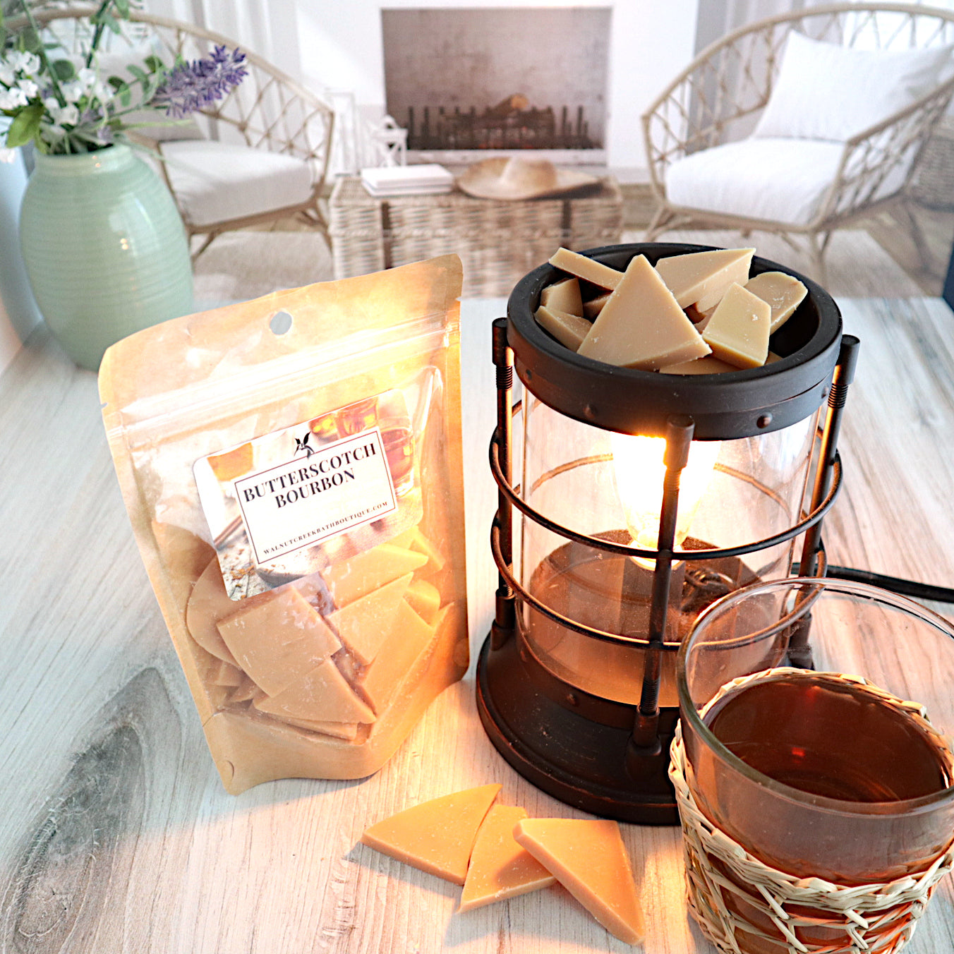 A butterscotch bourbon wax melt bag is standing next to a lit bronze wax melt burner. this has the tan bits of wax in it and there are some more of these scattered along the base. in the background is a fireplace flanked by 2 wicker chairs. there is a glass of what appears bourbon next to the wax burner