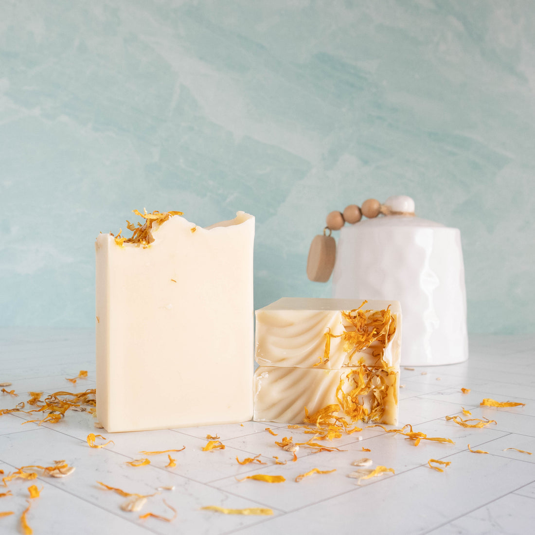 a bar of our soothing calendula soap is standing showing the creamy white soap. No swirls with this one, just creamy goodness. there are two other bars laying flat showing the tops that have some texture and then some calendula petals to give interest. there is a white crock in the background, a green wall in the background and on the surface are scattered calendula petals.
