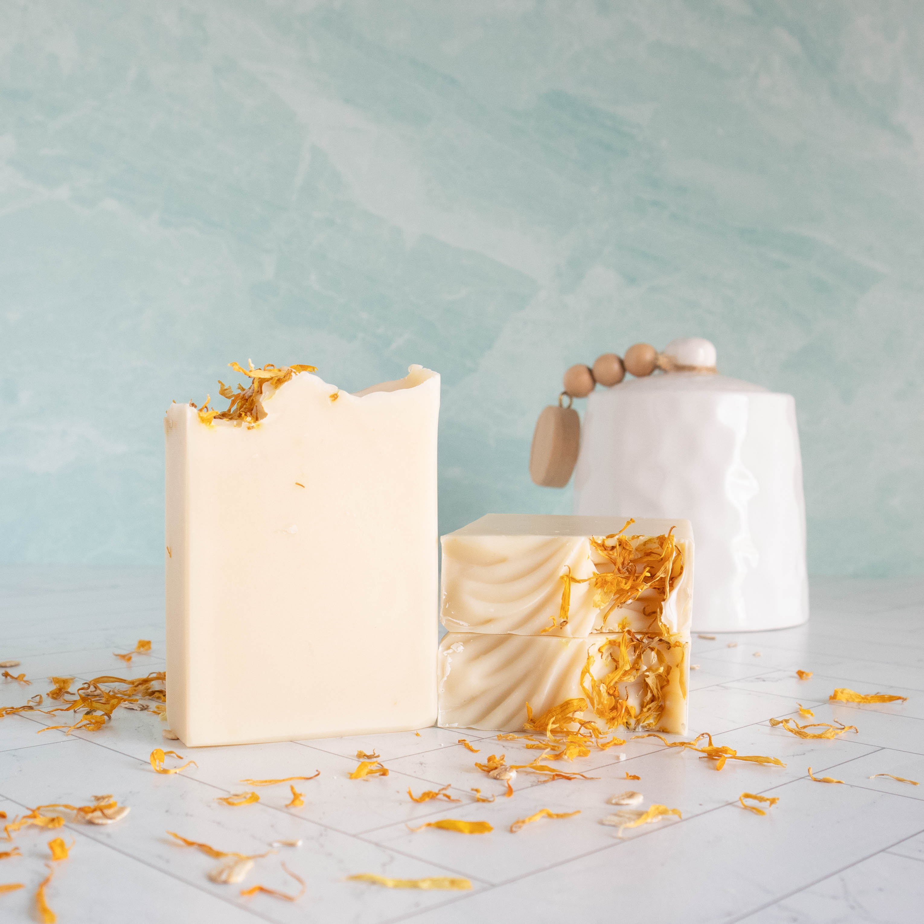 a bar of our soothing calendula soap is standing showing the creamy white soap. No swirls with this one, just creamy goodness. there are two other bars laying flat showing the tops that have some texture and then some calendula petals to give interest. there is a white crock in the background, a green wall in the background and on the surface are scattered calendula petals.