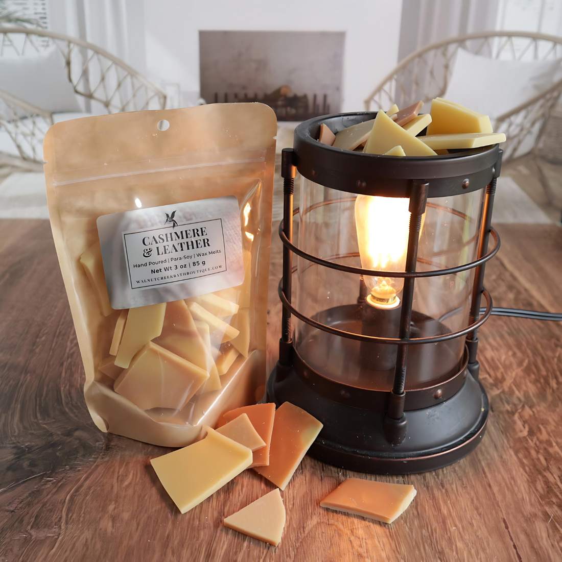 A cashmere and leather wax melt bag is standing next to a lit bronze wax melt burner. this has the tan bits of wax in it and there are some more of these scattered along the base. in the background is a fireplace flanked by 2 wicker chairs.