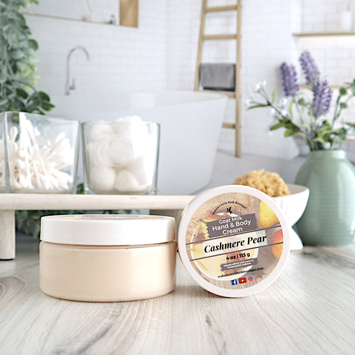 a large cashmere pear cream is sitting in front of a wood shelf that has q tips and cotton balls in glass dishes. there is a 4 ounce cashmere pear cream  on end next to the closed cream, to show the label. there is a white dish with a loofah in the background along with a sage green vase of flowers. there is an image of a bathroom in the backroom.