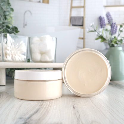 a large cashmere pear cream is sitting in front of a wood shelf that has q tips and cotton balls in glass dishes. there is another lagare cashmere pear cream on end next to the closed cream open, to show the cream on the inside. there is a white dish with a loofah in the background along with a sage green vase of flowers. there is an image of a bathroom in the backroom.