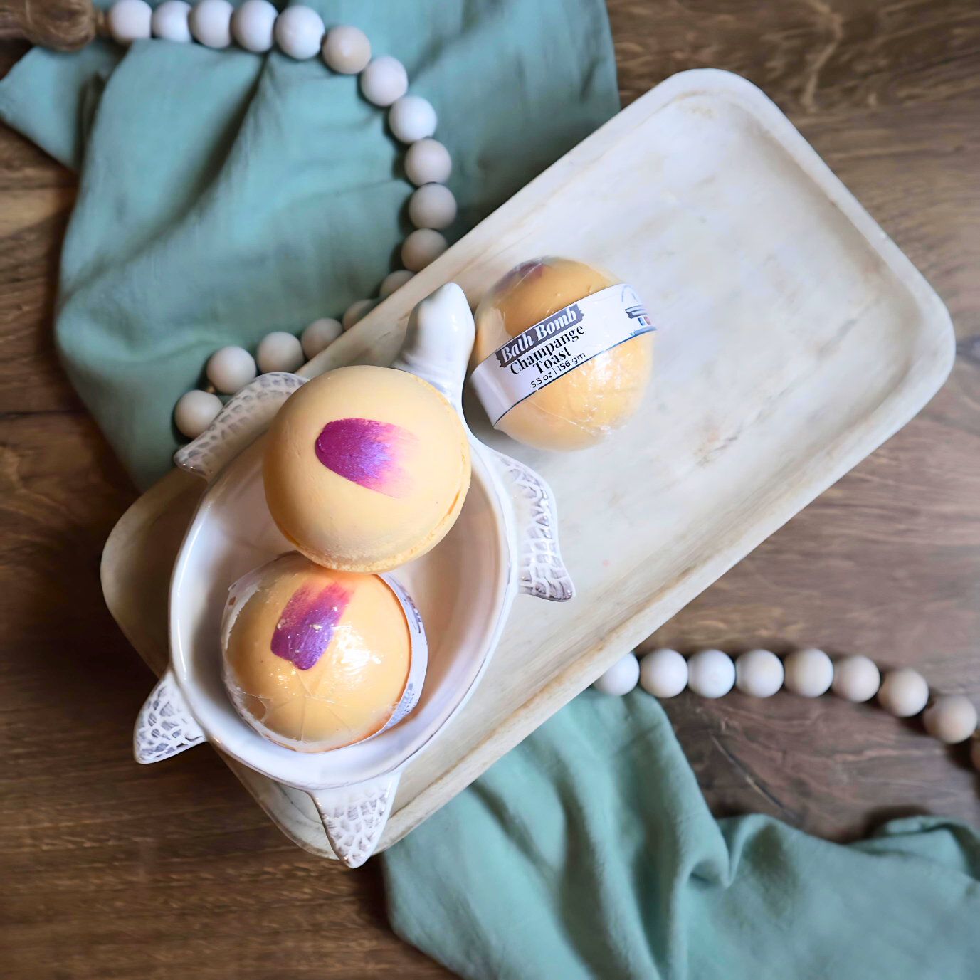 3 champagne toast bath bombs are shown sitting on a tray. two are in a soap dish in the shape of a turtle. the bath bombs are yellow orange in color with rosy purply swashes on the tops.  there is a warm wood base and a green towel with white beads running under the tray and through the picture.