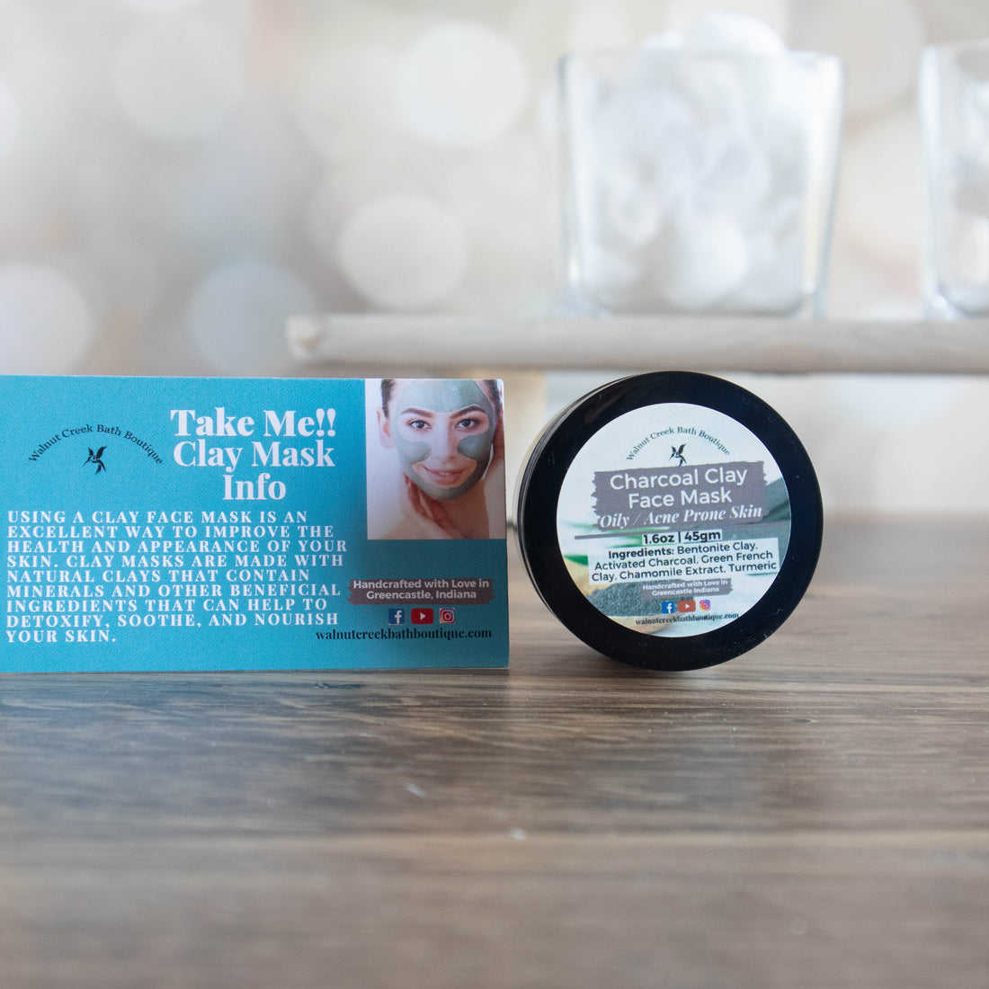 charcoal clay mask jar is sitting on its side so the label if facing our. sitting next to it is a clay mask card that comes in every order. these are sitting on a wood surface with a tray in the background that is holding a jar of cotton balls.