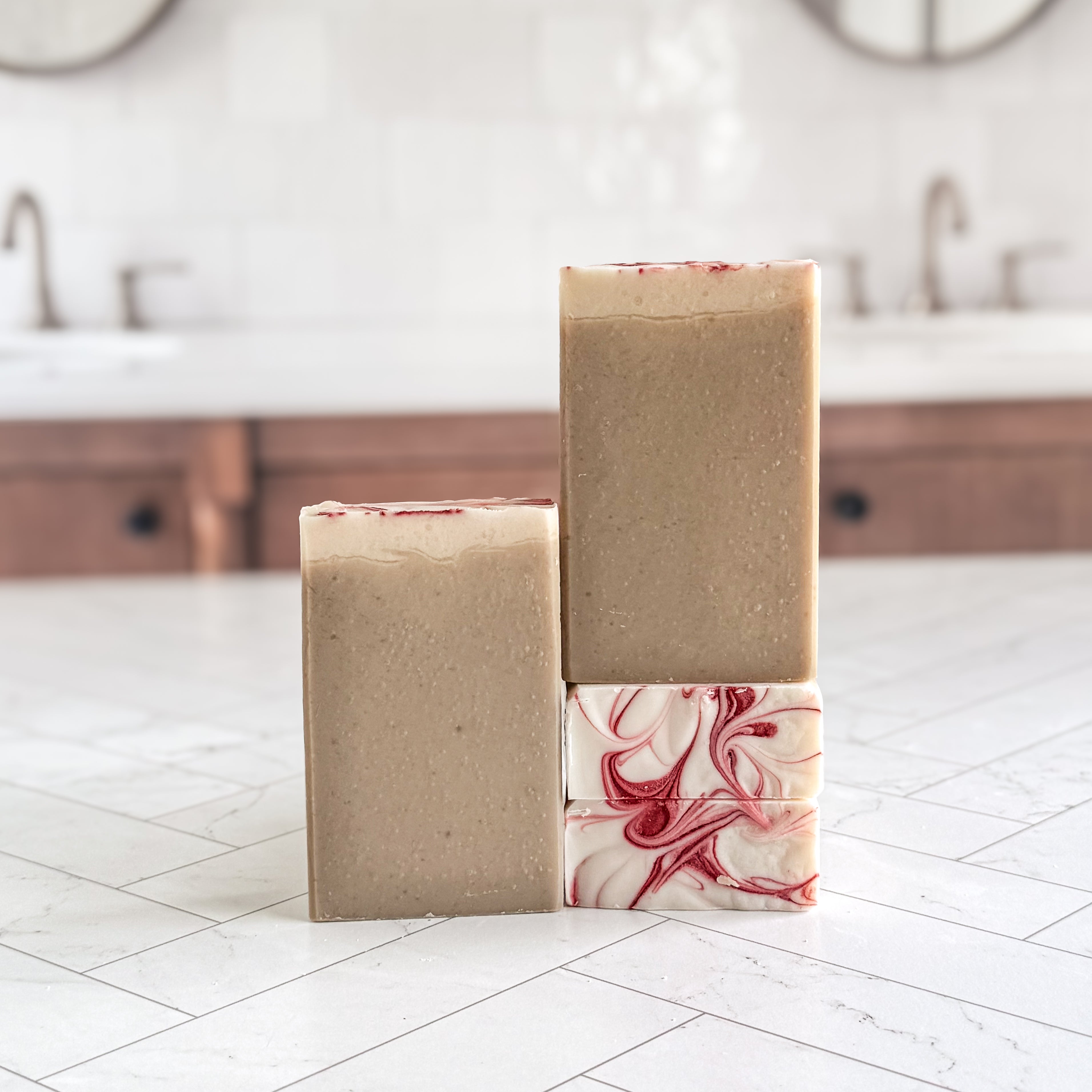 Three handmade soap bars displayed on a white marble countertop in a blurred bathroom setting. Two bars are a uniform light tan/beige color, and one bar features a white base with red marbled swirls.