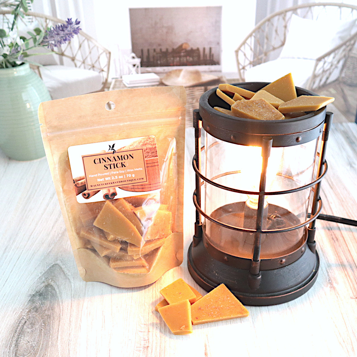 A cinnamon stick wax melt bag is standing next to a lit bronze wax melt burner. this has the brown bits of wax in it and there are some more of these scattered along the base.  in the background is a fireplace flanked by 2 wicker chairs. 