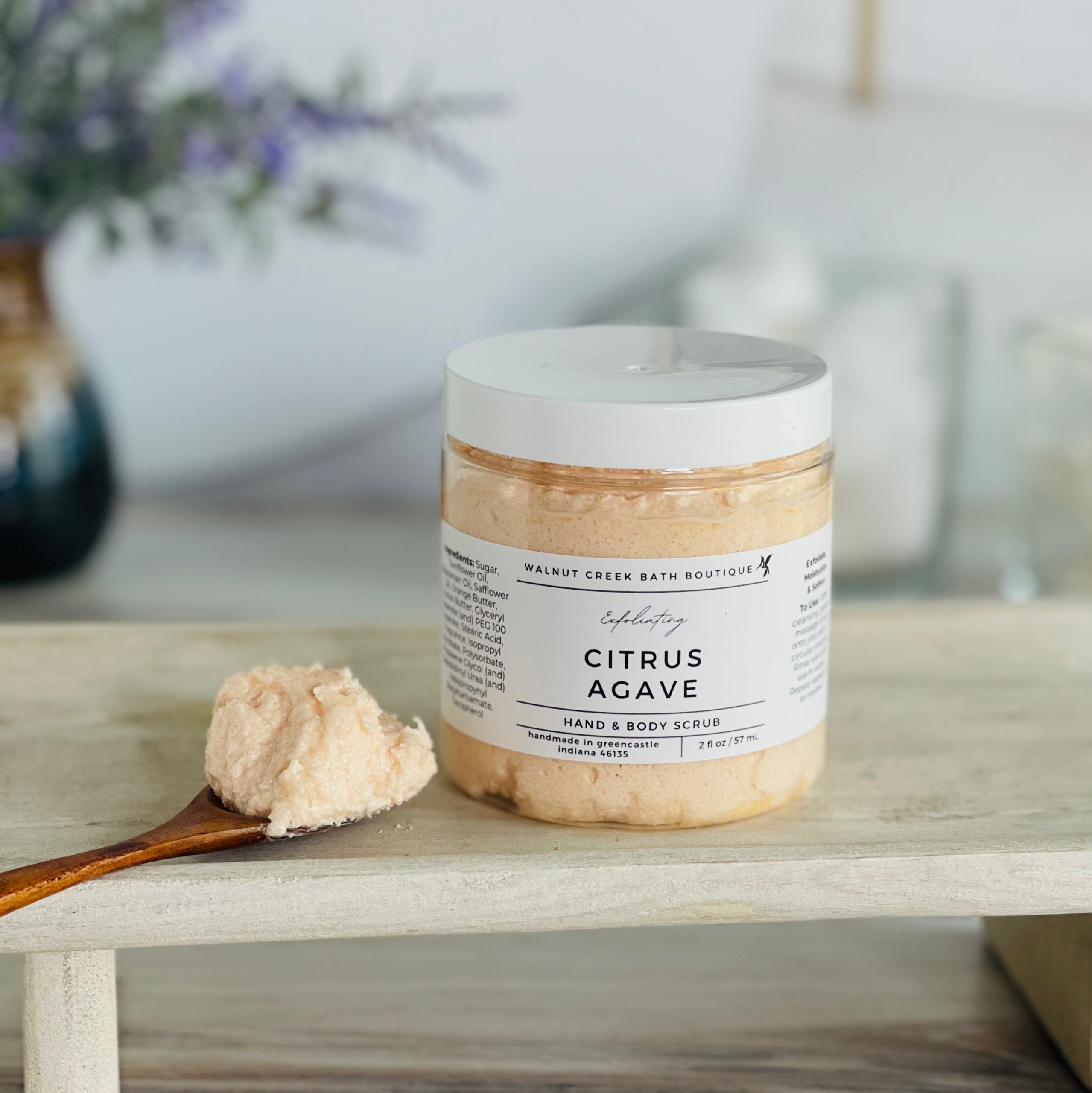 citrus agave scrub is shown on a tray with a wooden spoon mounded with the luscious scrub to show this fluffy consistency.. there is some lavender in the background to give interest to this very calm and neutral image to evoke a peaceful experience. 