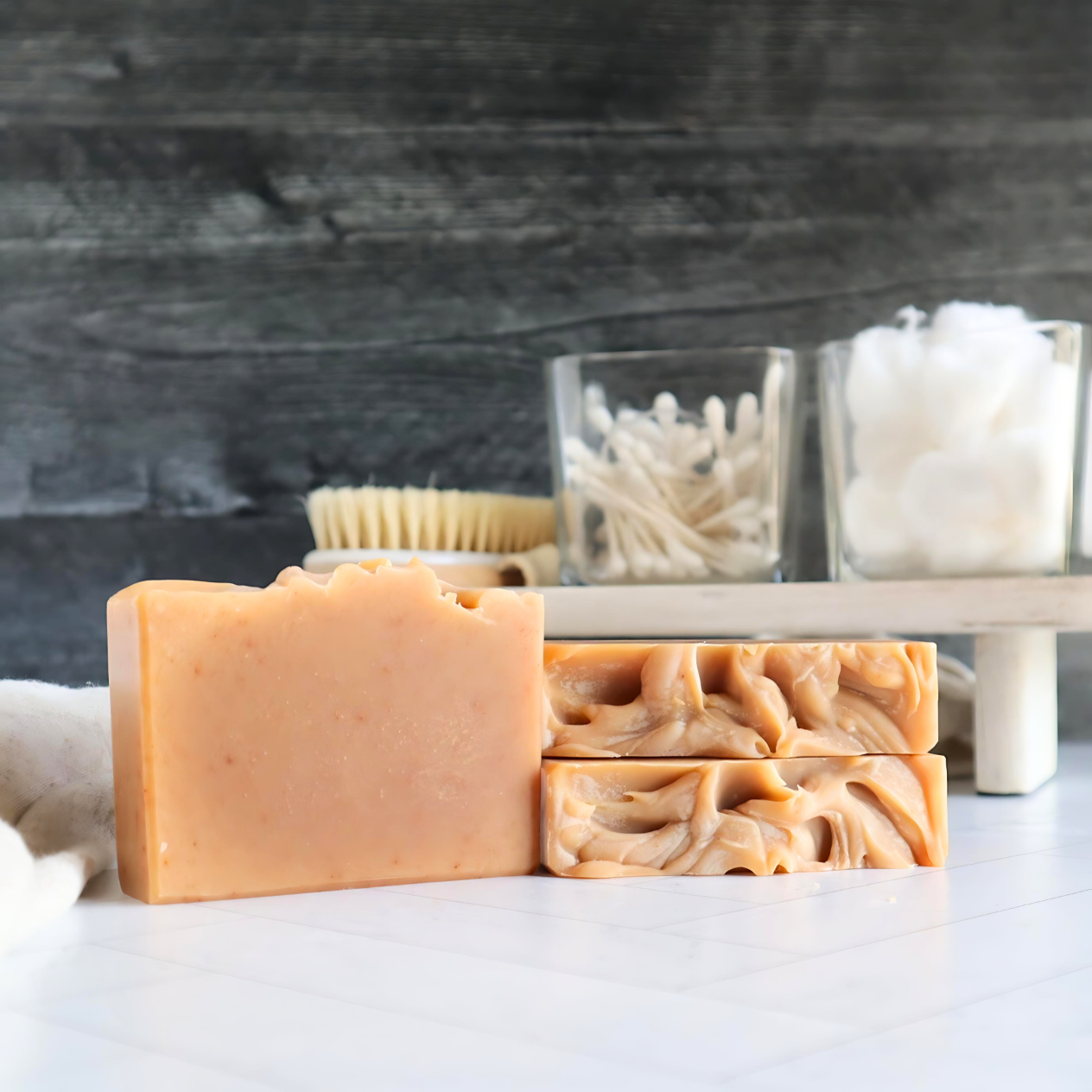 3 citrus burst soaps are shown, one is standing straight on with the front facing showing the orange color. the other two are laying flat to show a very textured top. in the background is a tray with glass dishes of cotton and q-tips.