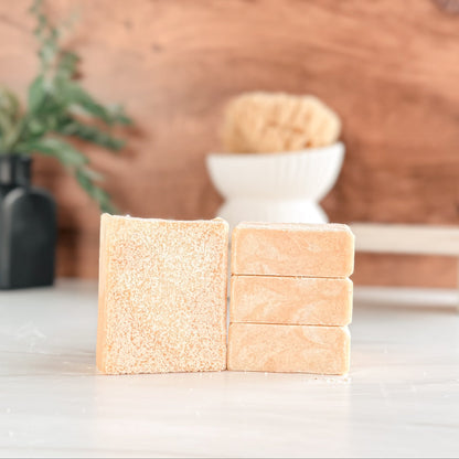 Three rectangular beige soap bars stacked on a white surface with a wooden background.