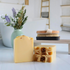 3 citrus serenity tallow soaps are shown with one standing tall facing you to show the creamy yellow color, this is standing next to 2 others that are laying flat to show the textured top with orange granules sprinkled along the middle. in the background is a try with a soap dish holding a bath brush.