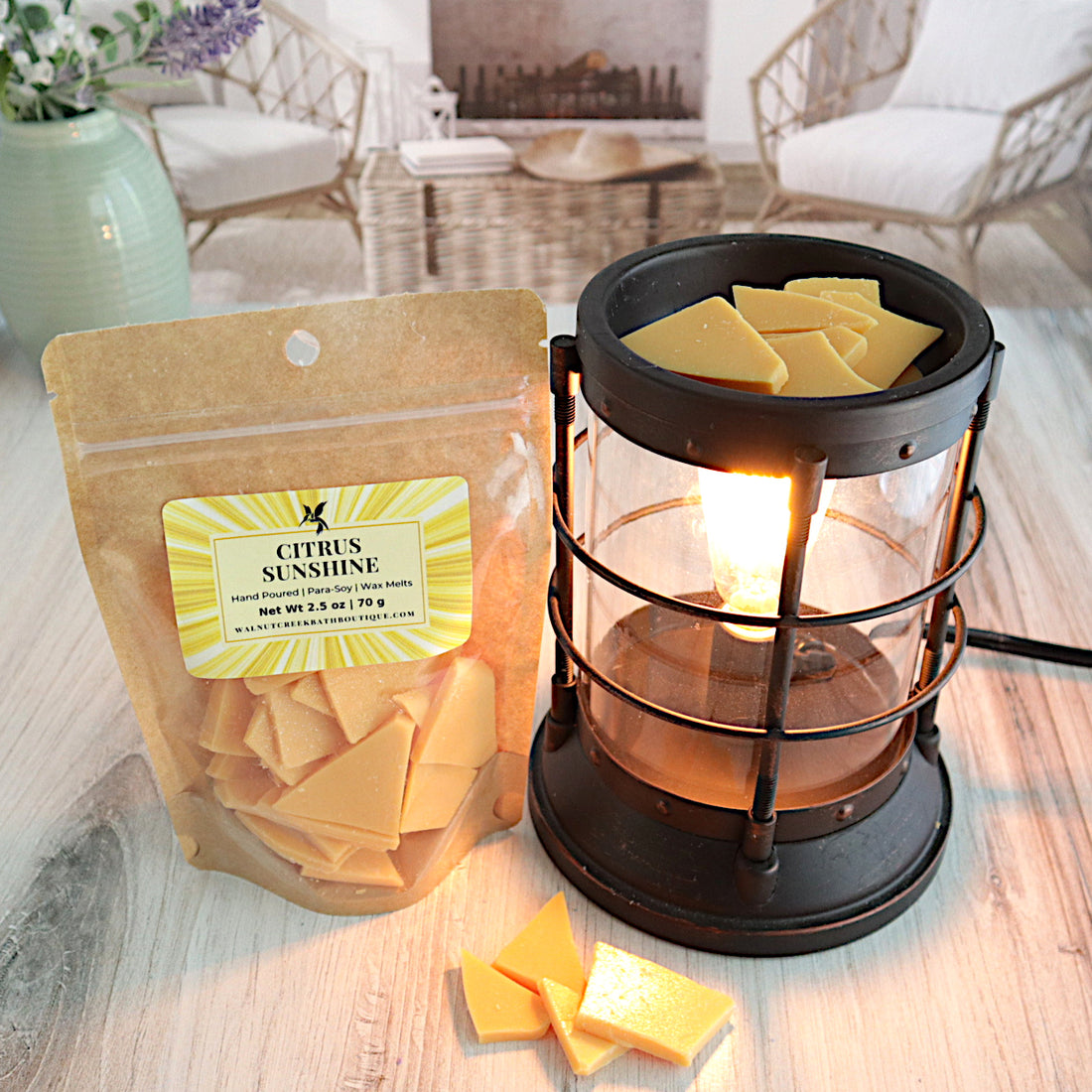 citrus sunshine is a pretty orange sparkly wax melt shown here in a bag with a fun sunshine beam label.  it is standing next to a lit burner full of the shard type pieces of wax. along the bottom of the burner are more pieces strewn about. this is all sitting on a washed out wooden base with a couple of chairs in the background next to a fireplace