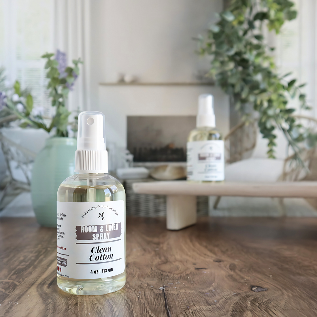 clean cotton room spray is shown . in the background is a second bottle on a tray next to a green vase with small flower buds. this is in front an image of a fireplace flanked by two chairs. there is some greenery hanging from the back right 