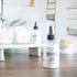 a clean cotton room spray is front and center. it is in a squat round bottle with black sprayer. in the background is a tray with some cotton balls and q-tips in jars along with another room spray. in the background is an image of a spa bathroom and a dish with a loofah