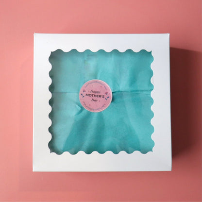 Closed white box with a window showing a teal tissue paper and a round sticker with the words happy mothers day on it. there is a coral colored background