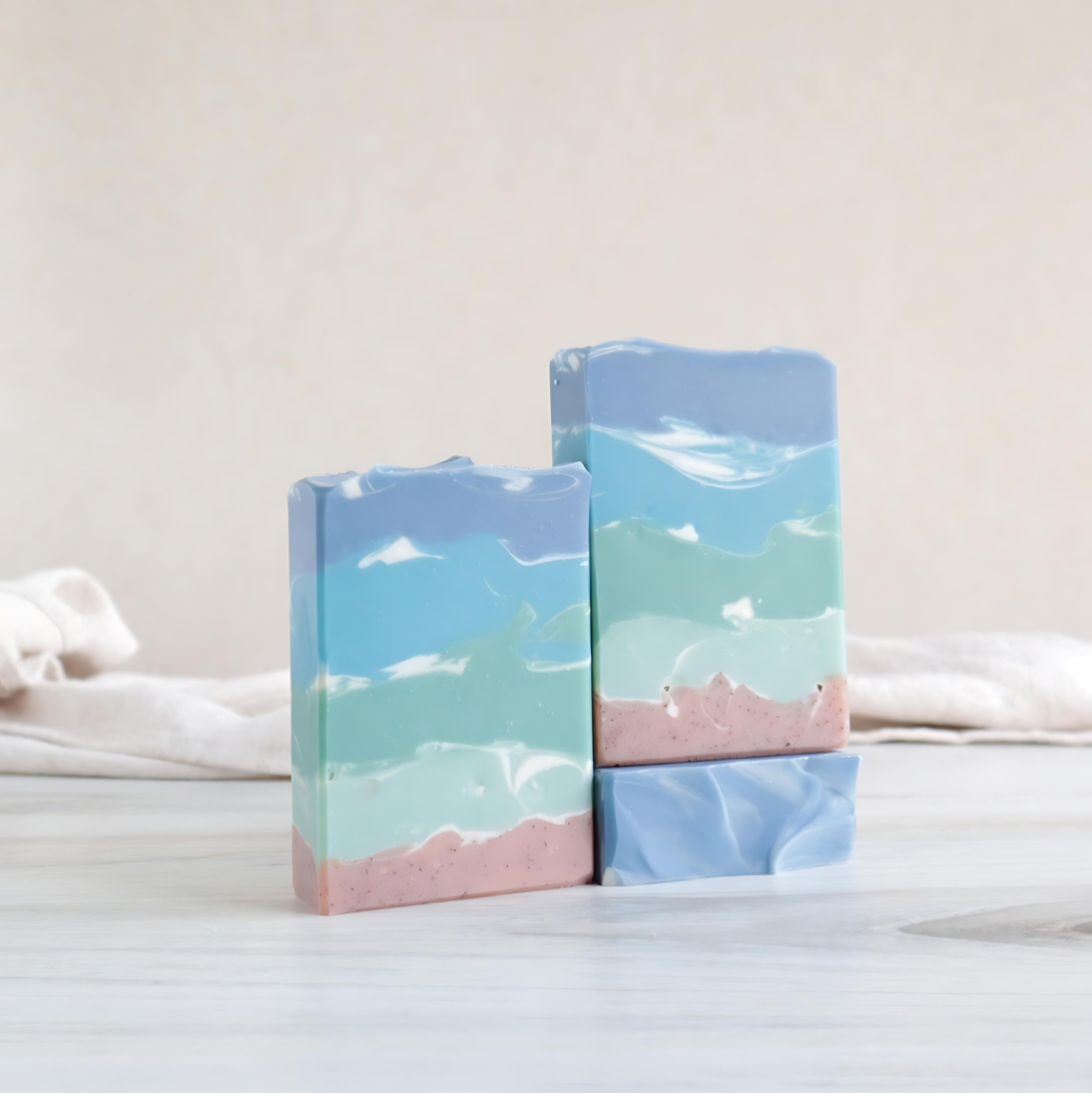 coastal air soaps are shown. 2 are standing tall facing out showing the  sandy colored bottom and then green soap with some white swirls going up and getting darker green as it goes all the way up to the top that is a smoky blue. there is a 3rd soap that is laying flat to show the textured top. there is a sandy colored background with a tan towel running behind the soaps.