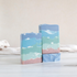 coastal air soaps are shown. 2 are standing tall facing out showing the  sandy colored bottom and then green soap with some white swirls going up and getting darker green as it goes all the way up to the top that is a smoky blue. there is a 3rd soap that is laying flat to show the textured top. there is a sandy colored background with a tan towel running behind the soaps.