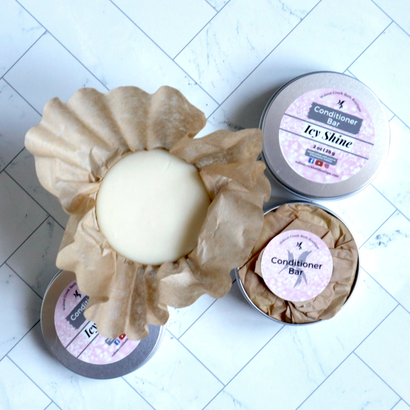 3 tins of conditioner bar are shown lying on a marble surface. One is closed showing the top label that is pink in color with the words conditioner bar and icy shine on top. another tin is open showing the inside packaging with brown paper wrapping and a pink sticker holding it closed. the sticker has a hummingbird behind the words conditioner bar written across it. the last tin is open with the paper open to show the conditioner bar in the tin.