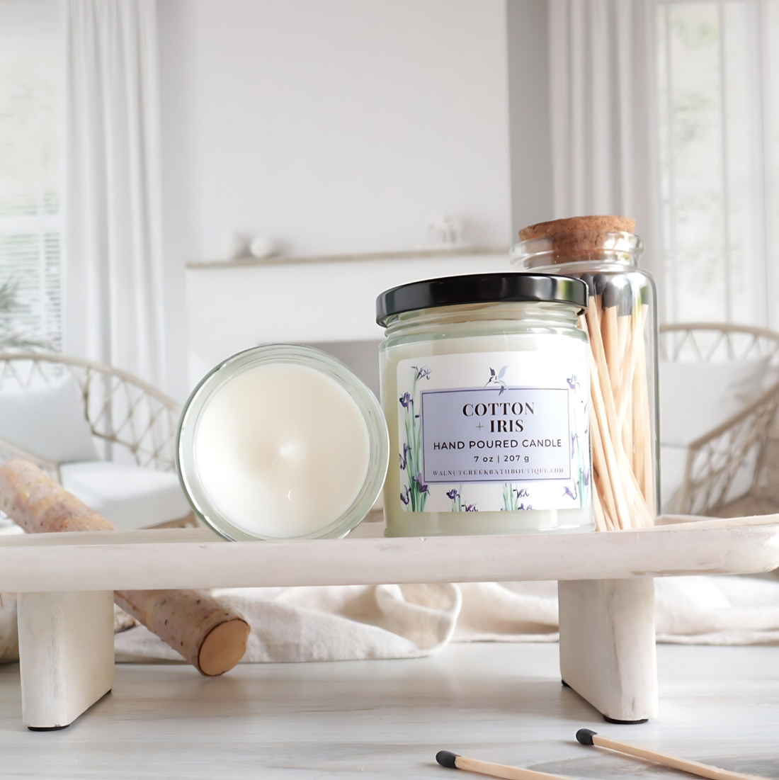 Cotton and Iris candle is show on a tray, one with the top off to show the creamy top, and next to that is a candle with the black lid and showing the label with irises in the background. there are matches scattered around and a glass jar of wooden matches behind the candle. in the background is an image of a fireplace with some logs in front.