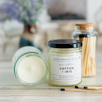 cotton and iris candle with lid to the right of another candle with the top off to show the creamy wax inside. there are a few matches scattered in front and a pretty glass jar of matches in the background