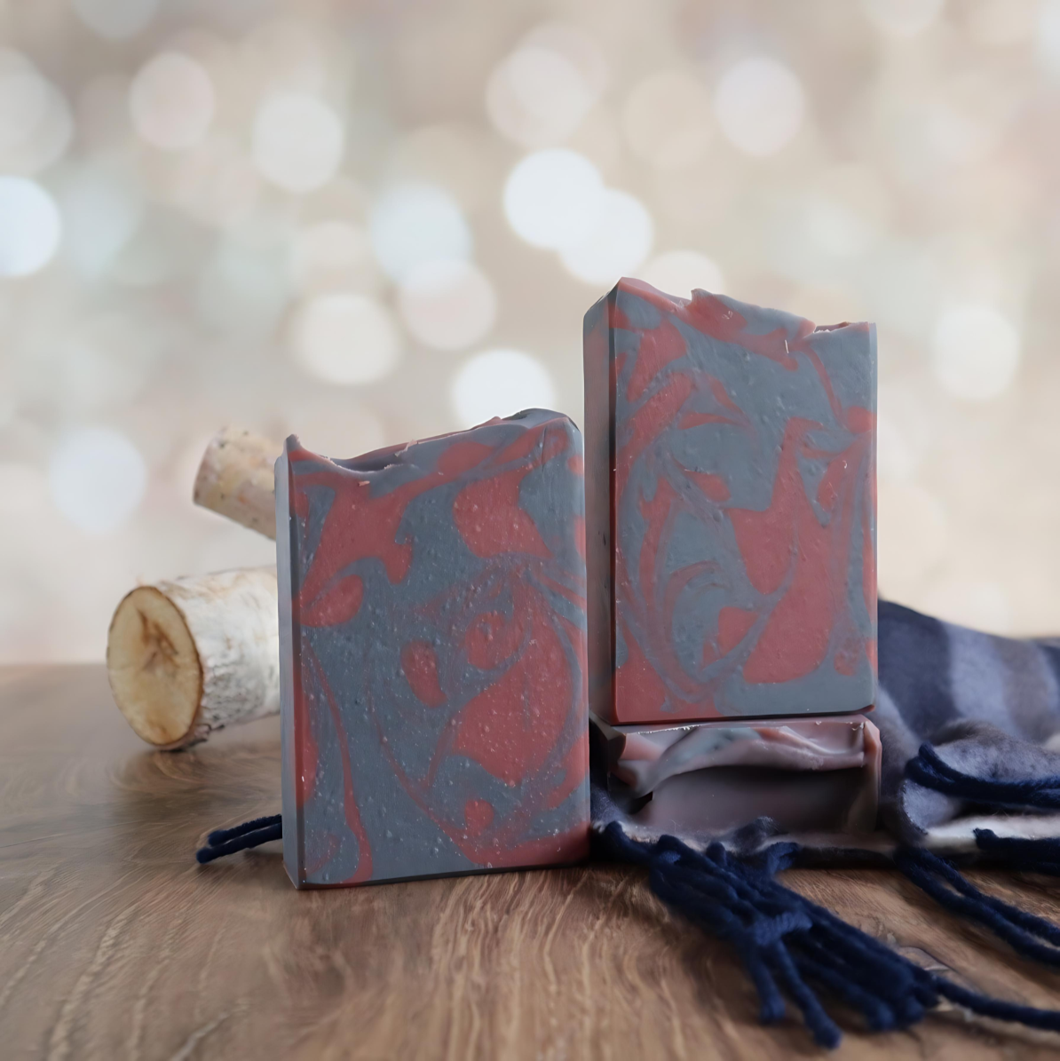 3 cozy flannel soaps are shown, one to the left and then another on the right that is standing on a 3rd that is laying flat to show the sculpted tops. the soaps are grey with a dark red swirl. behind the soaps are a couple of small logs and there is a blue flannel scarf in the back right. the background is an image of blurry twinkling lights.