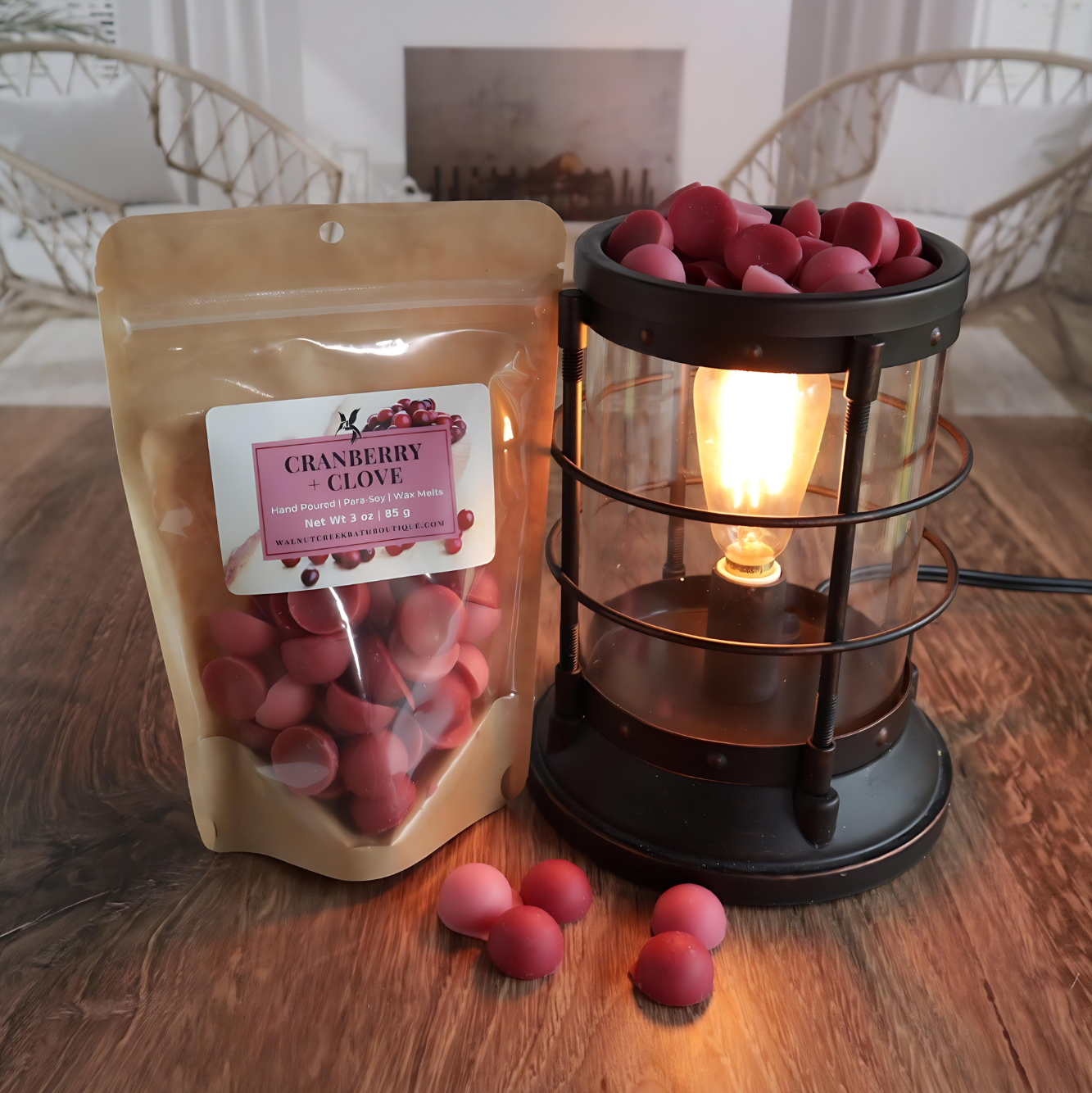 A cranberry and clove wax melt bag is standing next to a lit bronze wax melt burner. this has the red and pink bits of wax in it and there are some more of these scattered along the base. in the background is a fireplace flanked by 2 wicker chairs.