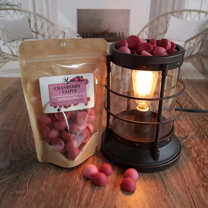 A cranberry and clove wax melt bag is standing next to a lit bronze wax melt burner. this has the red and pink bits of wax in it and there are some more of these scattered along the base. in the background is a fireplace flanked by 2 wicker chairs.