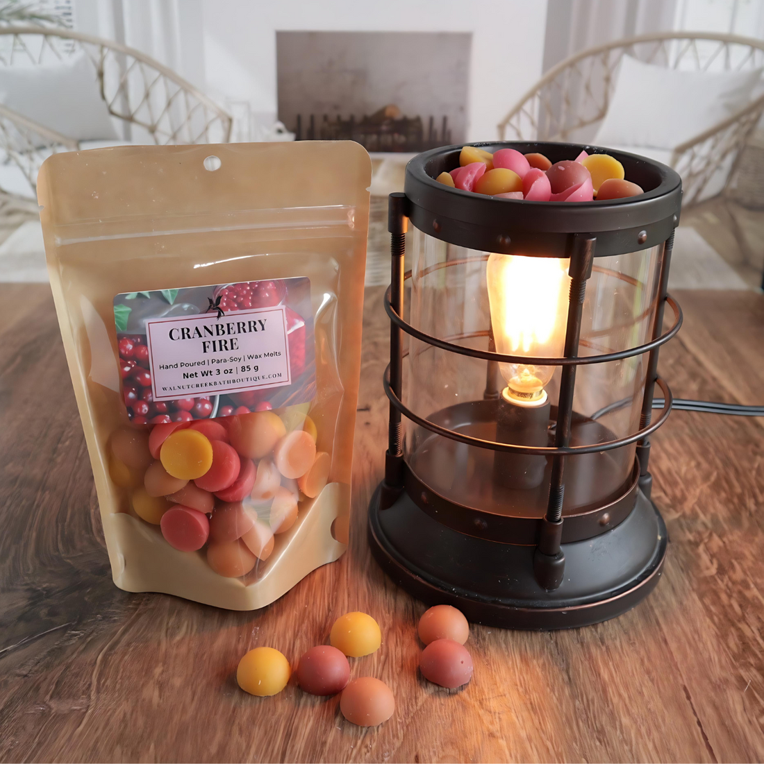 A cranberry fire wax melt bag is standing next to a lit bronze wax melt burner. this has the orange, yellow, brown and tanbits of wax in it and there are some more of these scattered along the base. in the background is a fireplace flanked by 2 wicker chairs.