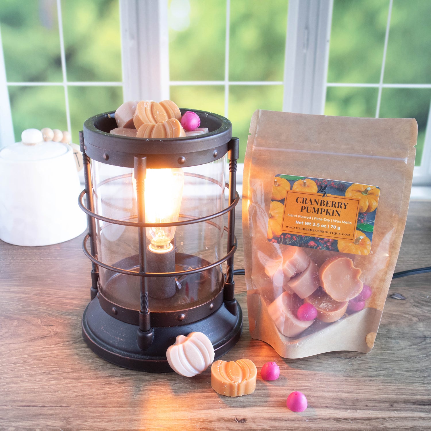 a cranberry pumpkin wax melt bag is standing to the right of a lit bronze burner. there are bits of wax in the shape of pumpkins and cranberries both in the burner and scattered along the base. there is a summer scene in the background through a window