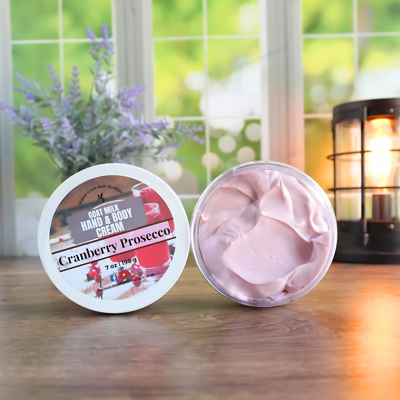 2 cranberry prosecco creams are shown standing on their sides. One to show the label and the other to show the creamy soft pink, swirly contents. in the back left is a vase with lavender buds and back right is a lit wax burner to add some light. behind all of this is a window with some greenery showing through.