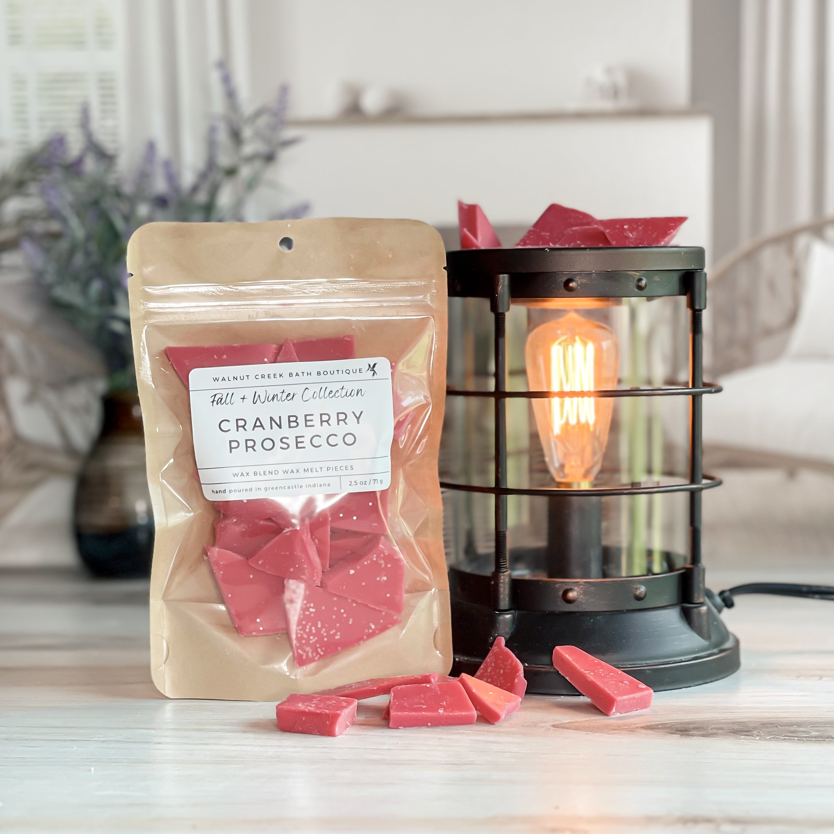 cranberry prosecco wax melt bag next to lit burner. there are wax melts on the table scattered about to shoe the pieces - these are red brittle pieces