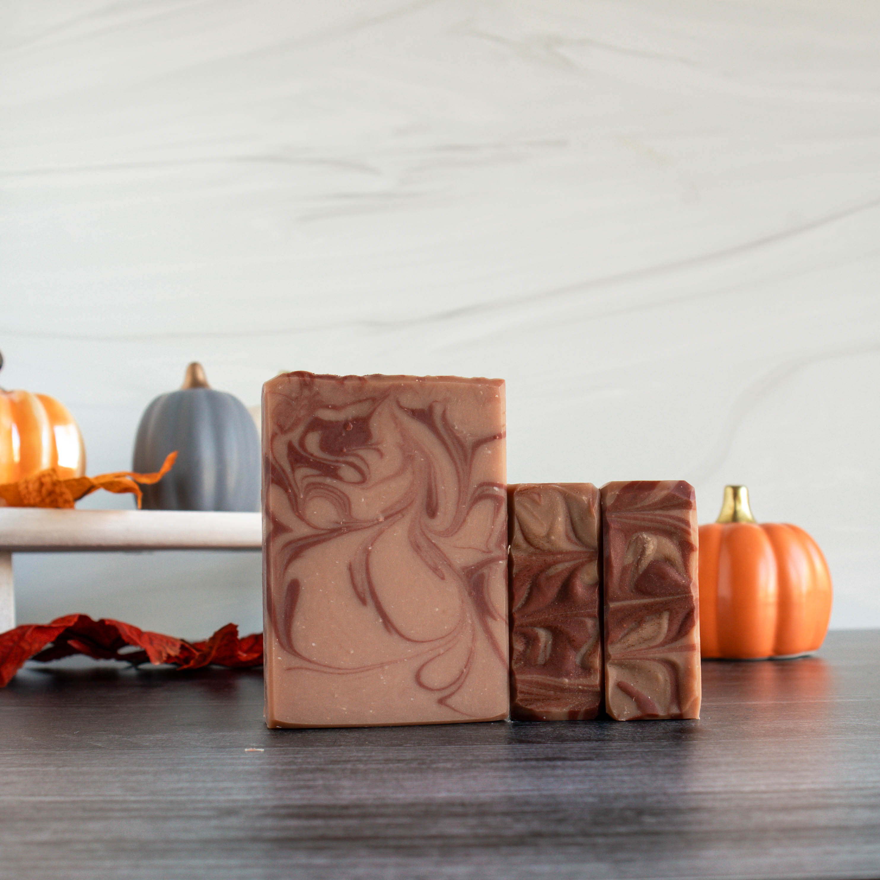 one cranberry pumpkin soap is standing showing the pretty tan and rust red swirls, 2 others are laying on the sides to show the top swirls in the same colors. there are several pumpkins in the background of different colors