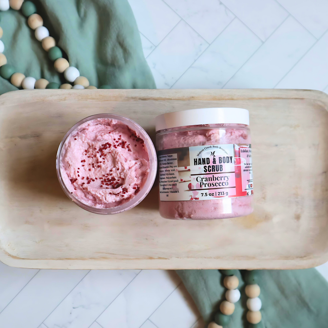 2 cranberry scrubs are sitting on a tray. the left scrub is without it tops to show the pretty pink scrub along with dark dots of cranberry seeds. the right scrub is laying flat to show the label.  underneath the tray is a green towel and some wooden beads.