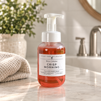 Walnut Creek Bath Boutique Crisp Morning Farrow Collection Tallow Hand Soap in a clear pump bottle with orange-amber liquid, set on a marble bathroom counter with a towel and plant in the background.
