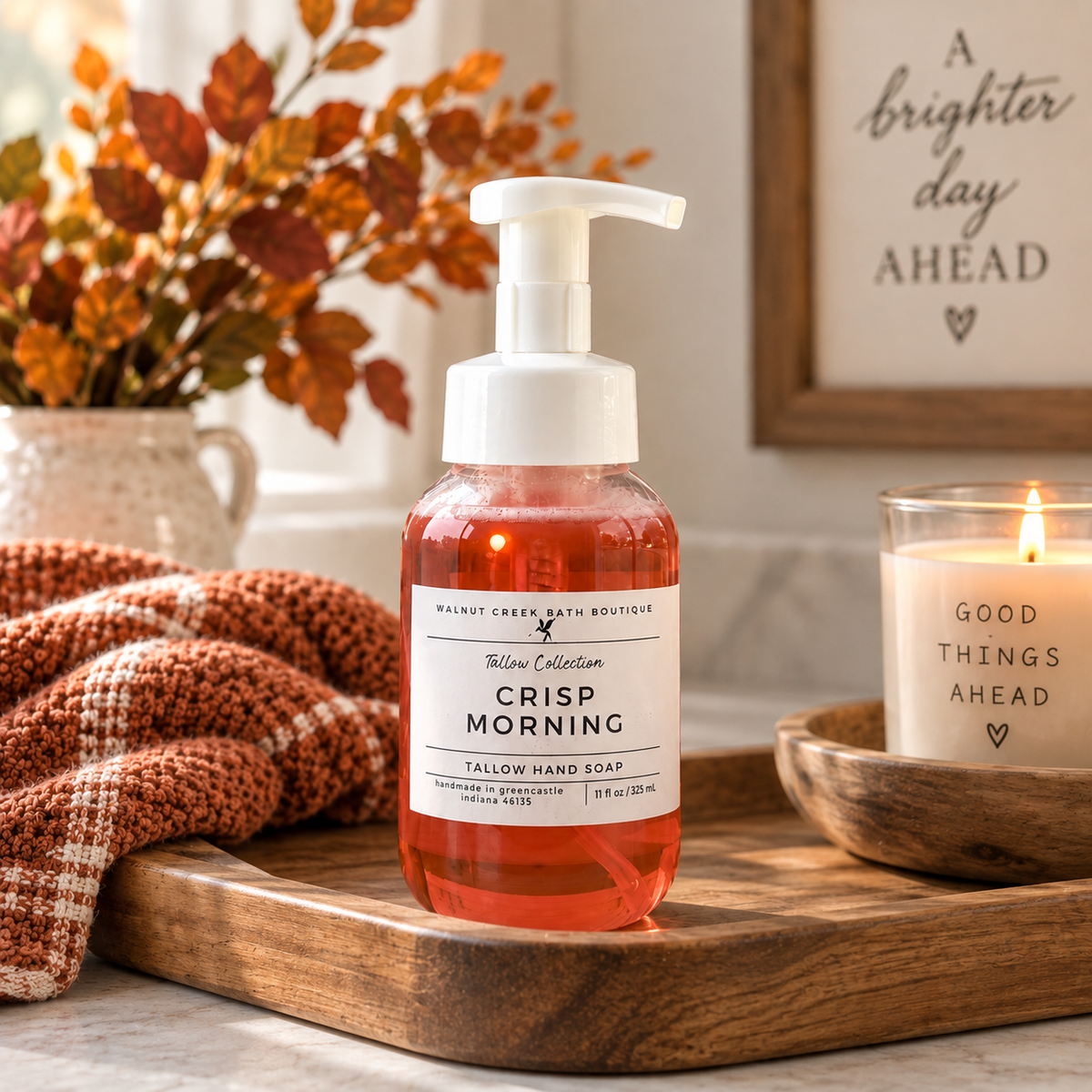 A pump bottle of Walnut Creek Bath Boutique Crisp Morning Tallow Hand Soap sits on a wooden tray in a cozy, styled bathroom setting with a lit candle, a folded rust-colored towel, and autumn foliage in the background.
