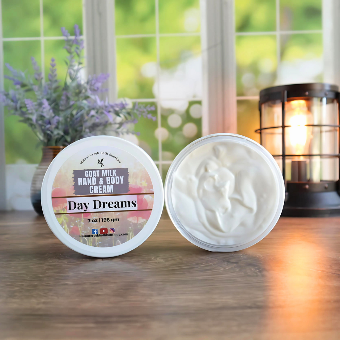 2 day dreams creams are shown standing on their sides. One to show the label and the other to show the creamy swirly contents. in the back left is a vase with lavender buds and back right is a lit wax burner to add some light. behind all of this is a window with some greenery showing through.