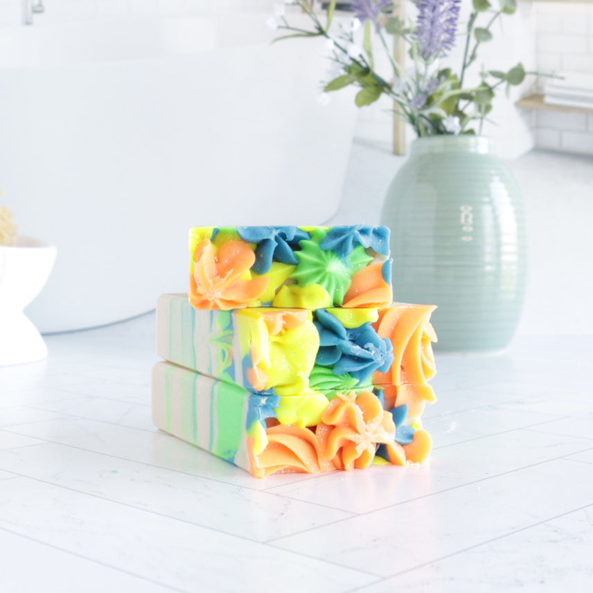 3 day dreams soaps are laying flat, stacked with the pretty tops forward to show the piping of rosettes, showing the bright green, blue, yellow and orange swirls. there is a vase in the background with some lavender buds peaking through.