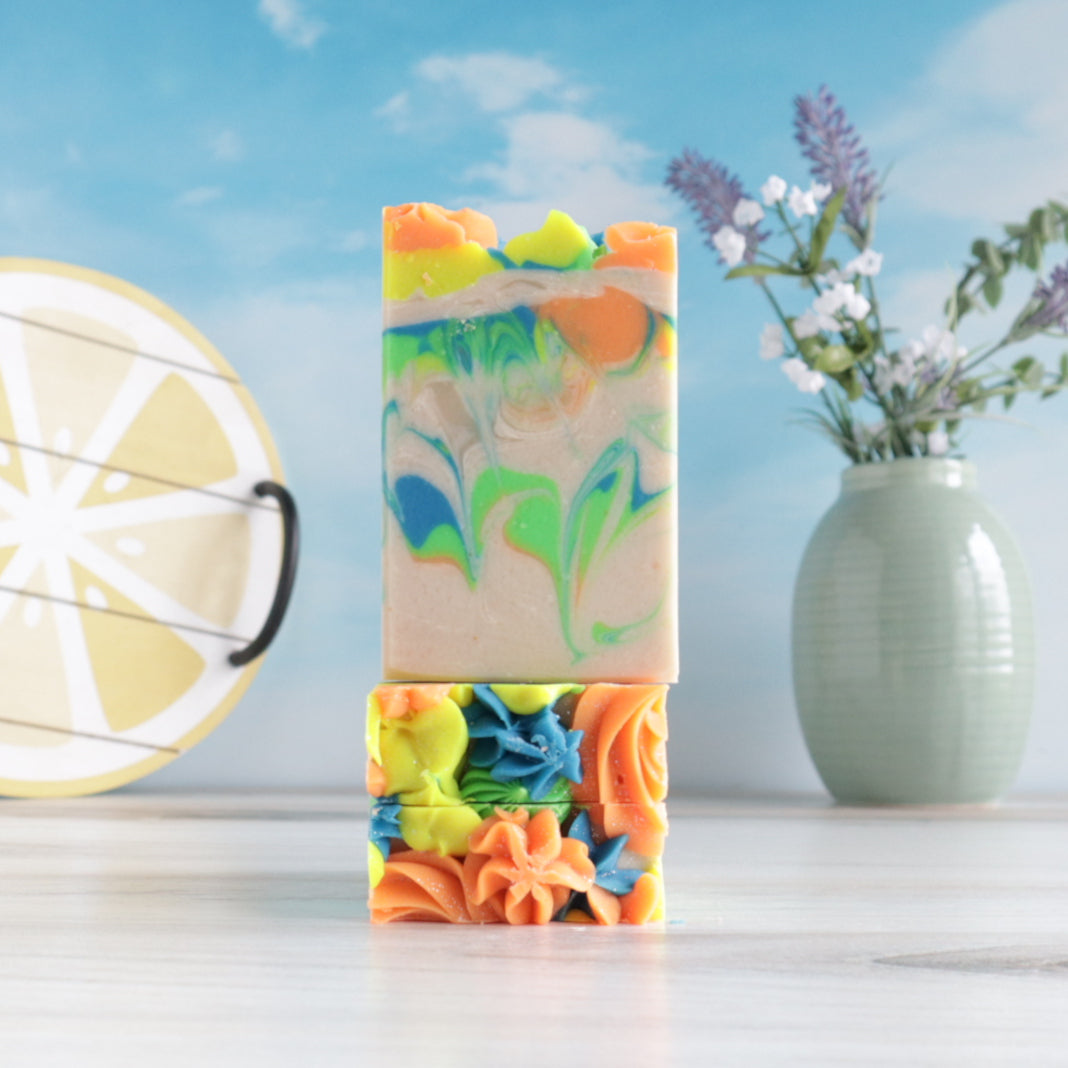 2 day dreams soaps laying flat, stacked with the pretty tops forward to show the piping of rosettes, there is another day dreams standing on top of them to show the bright green, blue, yellow and orange swirls. in the background is a pretty blue sky with a tray in the image of a lemon on one side and a vase with lavender and baby&