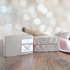 one dead sea mud soap is shown simply labeled and to the left of another two that is laying flat to show the tops of the soaps that have a texture to them. in the background is blurry twinkling lights and there is a tan towel along with a couple of small logs behind the soaps