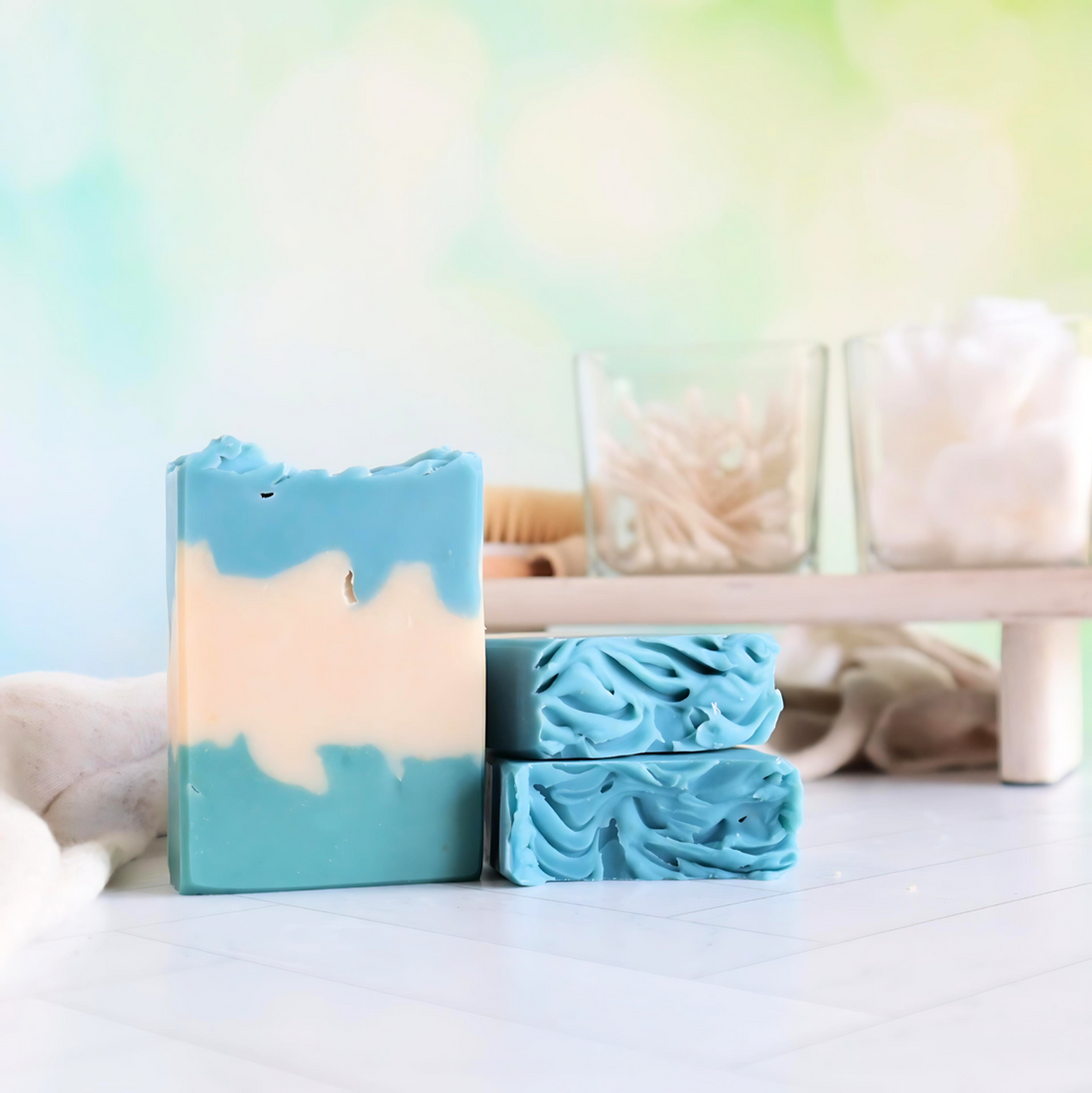 three eucalyptus and mint soaps are shown. One is standing facing  your to show the green bottom, cream middle and blue top of the design.  2 others are laying flat with the sculpted tops facing out. in the background is a tray with q-tips and cotton balls in glass containers. there is a light green backdrop.