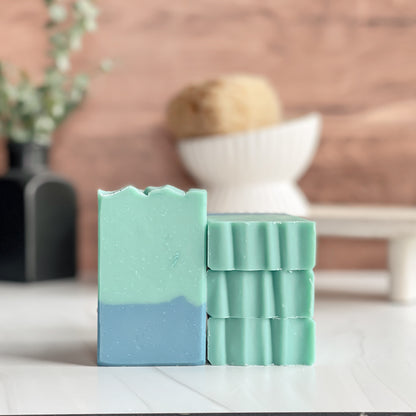 Two green and blue soap bars on a white surface with a blurred background