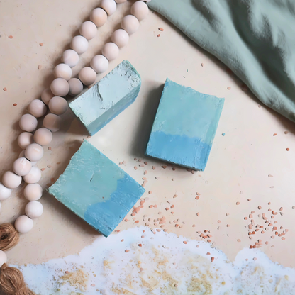 three flamingo beach soaps are shown, 1 standing tall on a sandy backdrop. the other 2 are laying flat with the image of the ocean just barely touching one. there are salts scattered around and a beach towel and wooden beads around the bars.