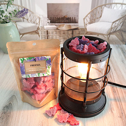 freesia wax melts are in the shape of cute little butterflies. the pieces are multi colored in shades of pink and purple. there is a bag of butterfly wax pieces standing next to a lit burner full of cuteness! there are some of the butterfly pieces scattered around the table in front of the burner.this is all sitting on a washed out wooden base with a couple of chairs in the background next to a fireplace