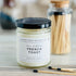 french toast candle with black lid. there are a few matches scattered in front and a pretty glass jar of matches in the background
