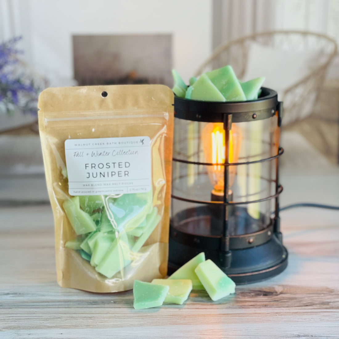 frosted juniper wax melt bag is full and standing next to a rustic farmhouse type burner that is on and filled with the green brittle pieces of wax. there are also a few shards of the brittle on the table in front of both to show the size and color of the pieces