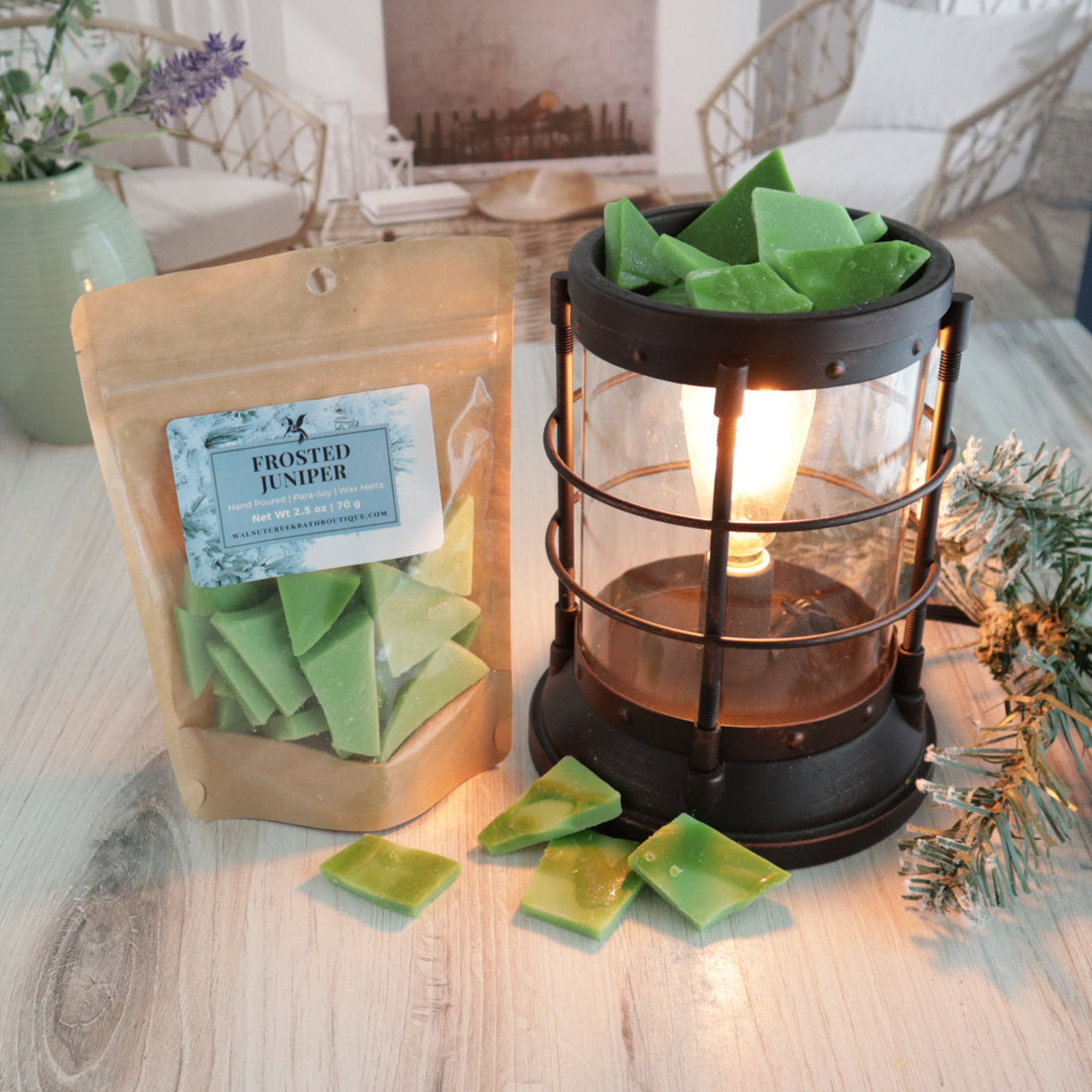 a bag of frosted juniper wax melts are standing next to a lit wax melt burner. this has some of the green wax pieces piled in it. there are also some pieces scattered around between the bag and the burner. there is a sprig of frosted pine branch to the right of the burner. this is all sitting on a washed out wooden base with a couple of chairs in the background next to a fireplace
